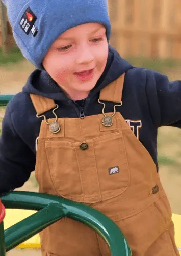 Young child in brown overalls and blue hat playing outdoors.