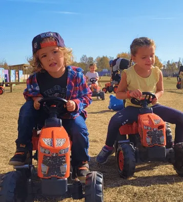 Two young children riding toy tractors outdoors on a sunny day.